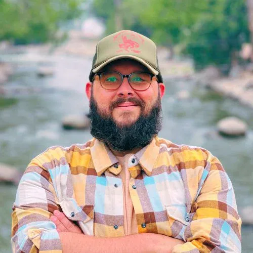Bearded man in cap and plaid shirt outdoors near river, representing church community leadership.
