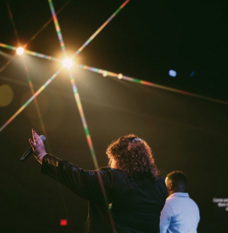 A worship leader with arms raised under bright stage lights during a church service